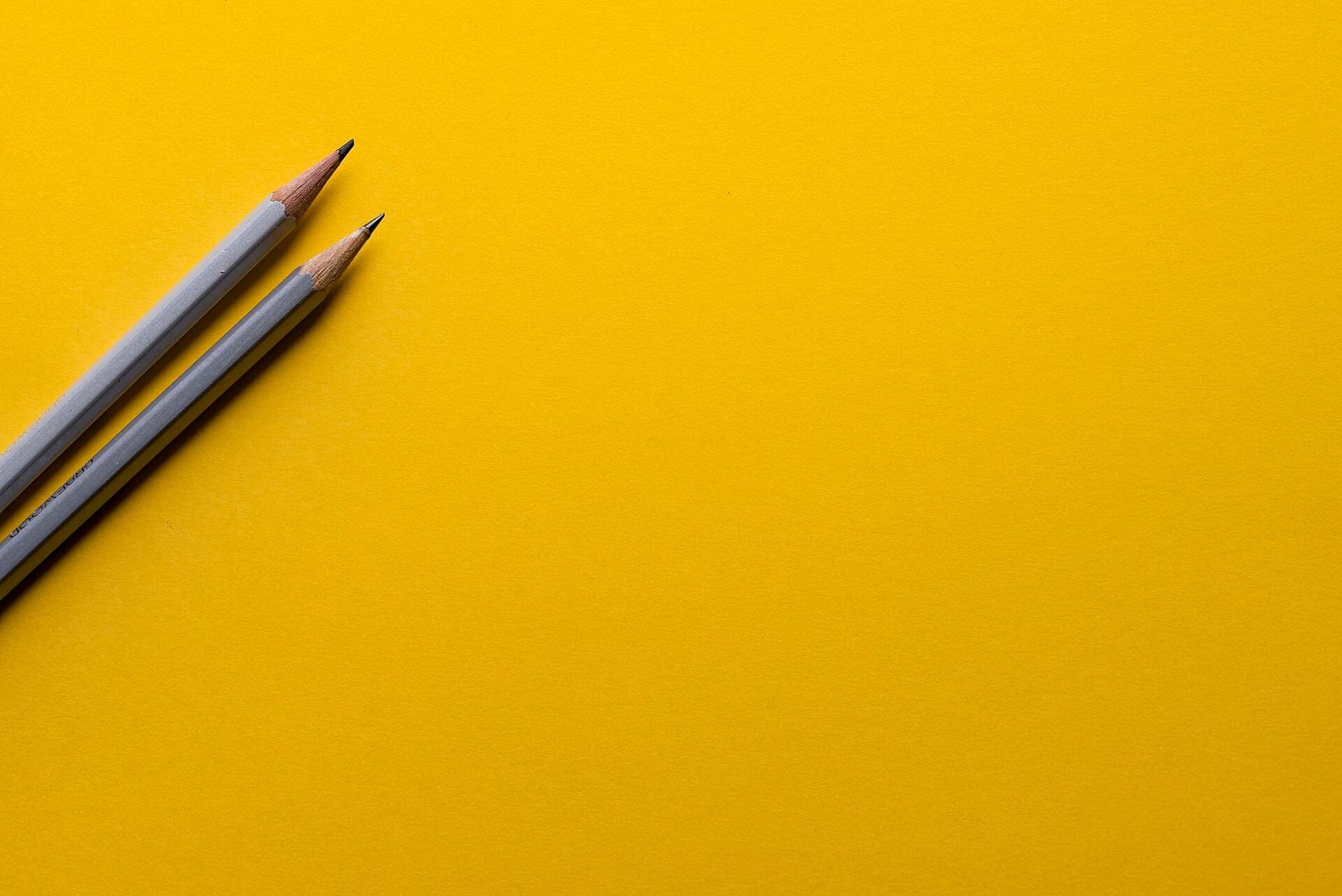 Two sharpened gray pencils lying diagonally in the corner of a bright yellow background.