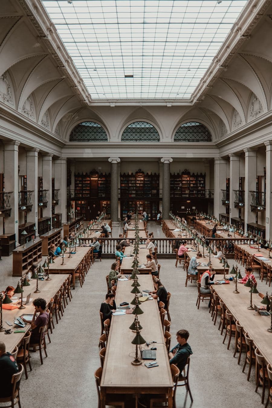A grand skylit library reading room with students working at long wooden tables.