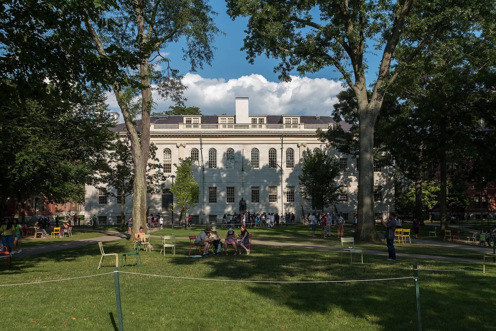 University Hall in Harvard Yard on a summer day, with visitors gathered by the John Harvard statue and people sitting on lawn chairs under tall trees.