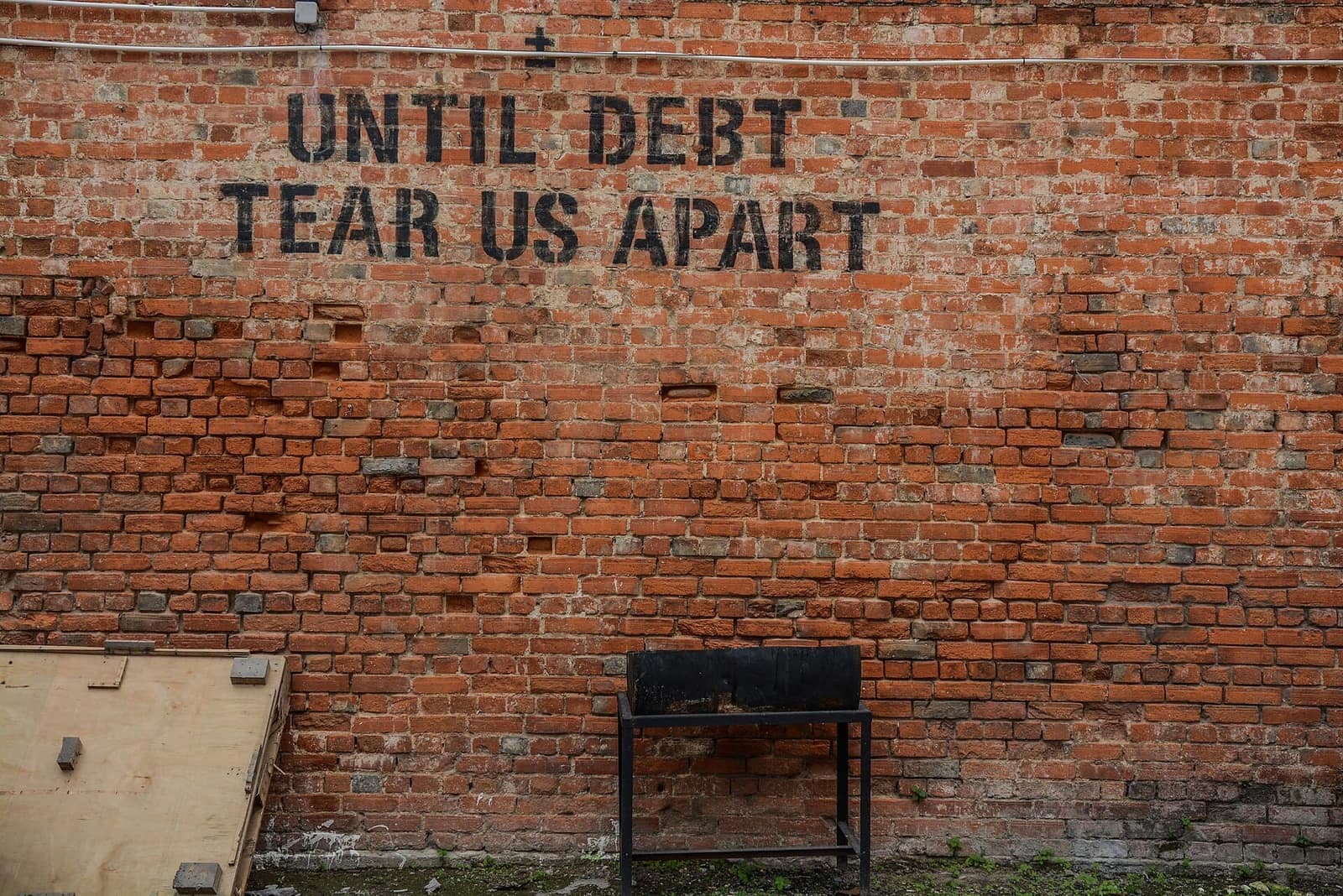 A red brick wall stenciled in black with the words "Until debt tear us apart" above an empty metal stand.